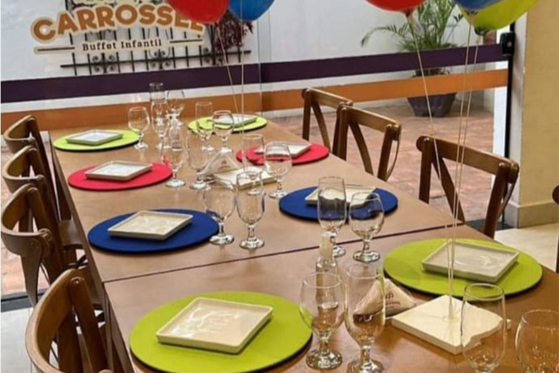 Mesa comprida posta com pratos coloridos, com o letreiro do Espaço Carrossel na parede